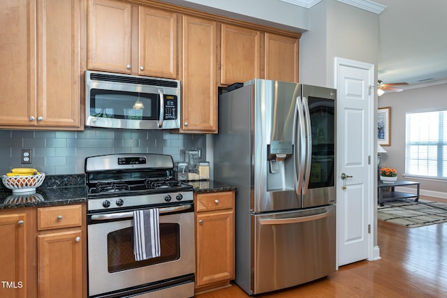 kitchen with appliances with stainless steel finishes, ornamental molding, light wood-type flooring, tasteful backsplash, and dark stone countertops