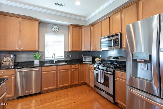 kitchen with stainless steel appliances, a sink, crown molding, dark stone countertops, and light wood-type flooring