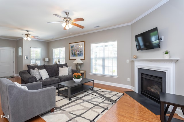 living room with light wood-type flooring, a ceiling fan, ornamental molding, baseboards, and a glass covered fireplace