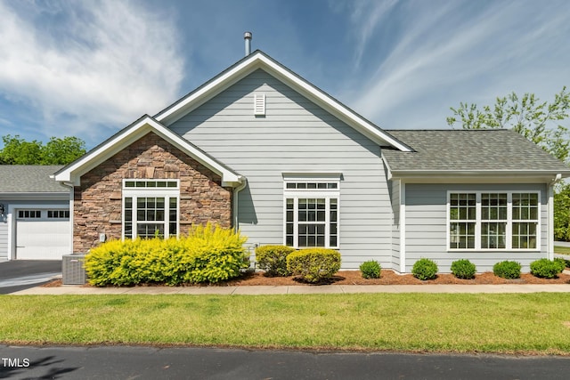view of front of property with an attached garage, a shingled roof, stone siding, a front lawn, and central air condition unit