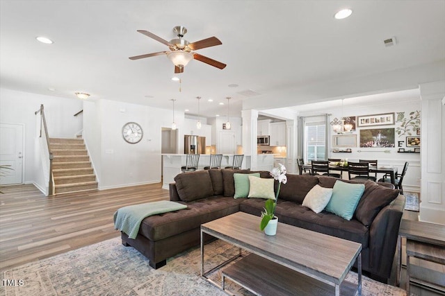 living room with visible vents, light wood-style floors, baseboards, stairway, and ceiling fan with notable chandelier
