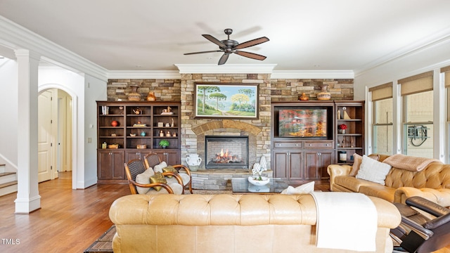 living room featuring a ceiling fan, a stone fireplace, wood finished floors, crown molding, and stairs