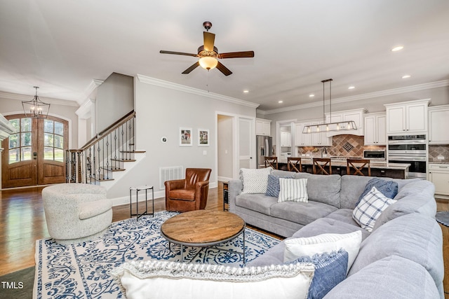 living area featuring ceiling fan with notable chandelier, visible vents, dark wood-style flooring, ornamental molding, and stairs
