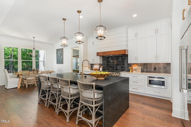 kitchen with dark countertops, a sink, custom range hood, tasteful backsplash, and a center island with sink