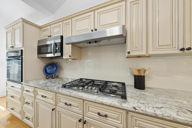 kitchen featuring decorative backsplash, light stone countertops, under cabinet range hood, stainless steel appliances, and cream cabinetry