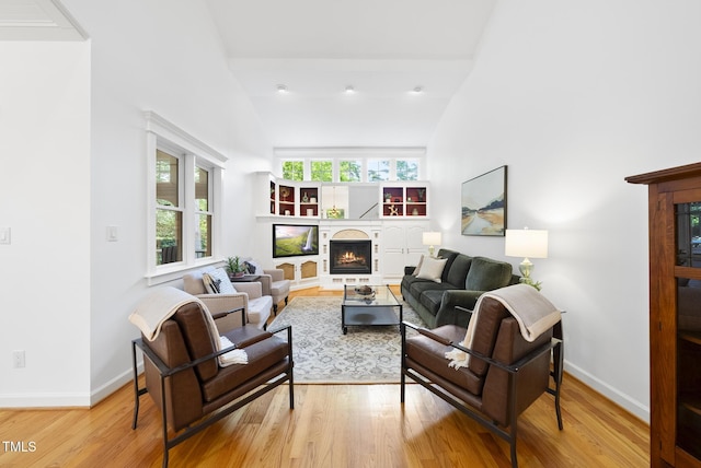 living room featuring high vaulted ceiling, baseboards, a lit fireplace, and light wood-style flooring