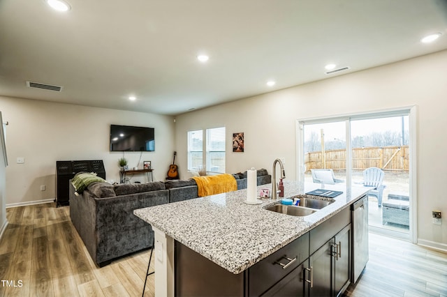 kitchen featuring stainless steel dishwasher, light stone counters, a sink, an island with sink, and light wood-style flooring