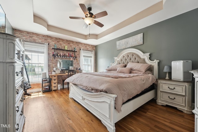 bedroom with wood finished floors, brick wall, a tray ceiling, and multiple windows