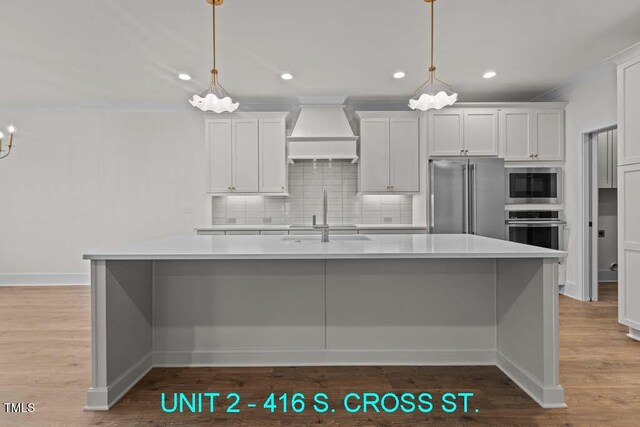 kitchen with appliances with stainless steel finishes, a sink, premium range hood, decorative backsplash, and a center island with sink