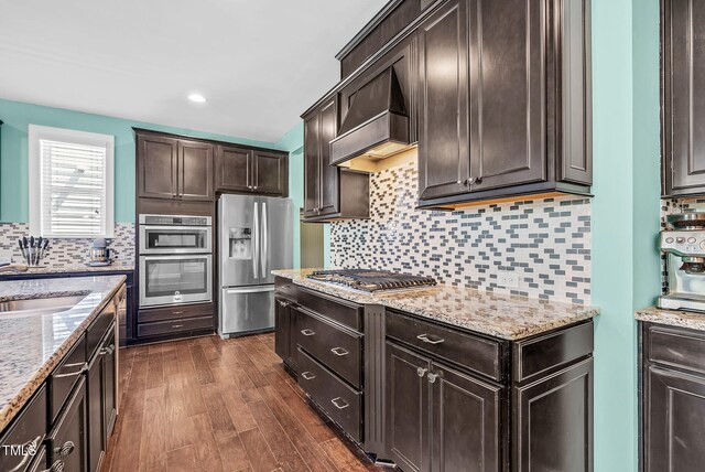 kitchen featuring custom range hood, appliances with stainless steel finishes, dark brown cabinets, dark wood-type flooring, and backsplash