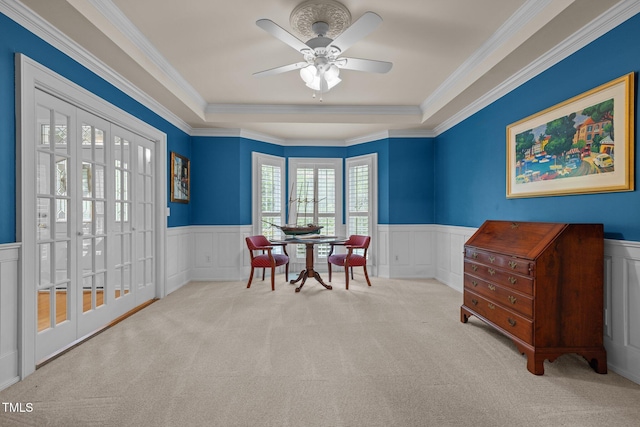 carpeted dining room featuring a wainscoted wall, a decorative wall, ornamental molding, a ceiling fan, and a tray ceiling