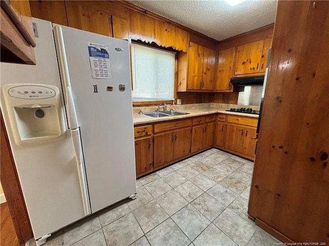 kitchen with a sink, a textured ceiling, white refrigerator with ice dispenser, brown cabinetry, and under cabinet range hood