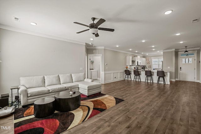 living room with dark wood-style floors, visible vents, a ceiling fan, and crown molding