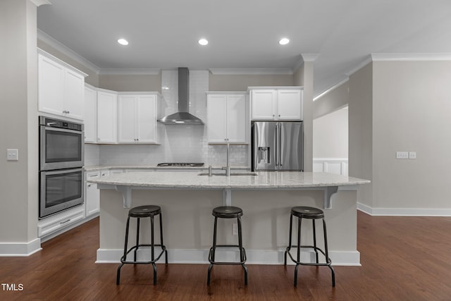 kitchen with stainless steel appliances, wall chimney range hood, ornamental molding, a sink, and white cabinetry