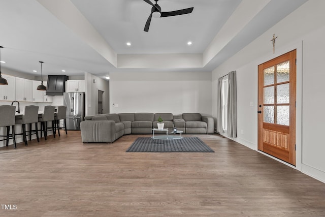 living room featuring ceiling fan, a raised ceiling, baseboards, light wood-style floors, and recessed lighting