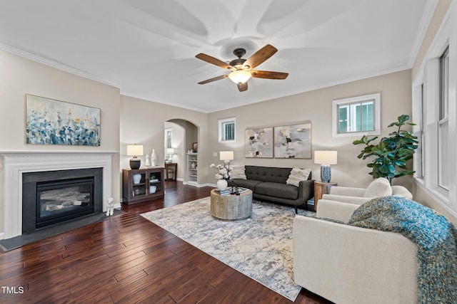 living area with arched walkways, a ceiling fan, dark wood-style floors, crown molding, and a fireplace with flush hearth