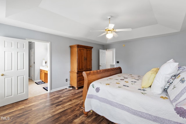 bedroom featuring a raised ceiling, wood finished floors, ensuite bath, baseboards, and ceiling fan