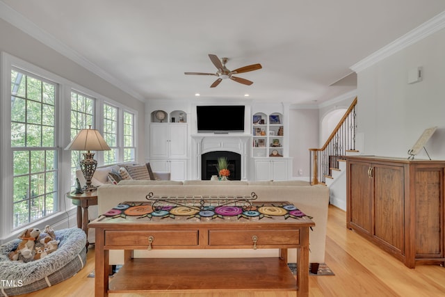 living room featuring a glass covered fireplace, ceiling fan, stairway, built in shelves, and light wood finished floors