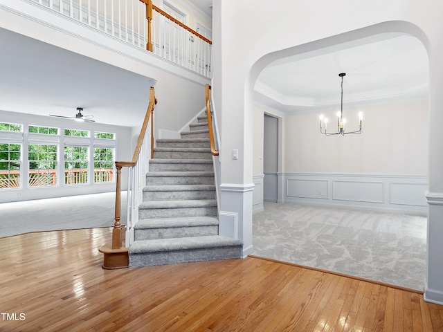 staircase with crown molding, arched walkways, a wainscoted wall, ceiling fan with notable chandelier, and hardwood / wood-style floors
