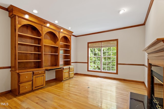 unfurnished office featuring crown molding, built in desk, light wood-style flooring, baseboards, and a fireplace with flush hearth