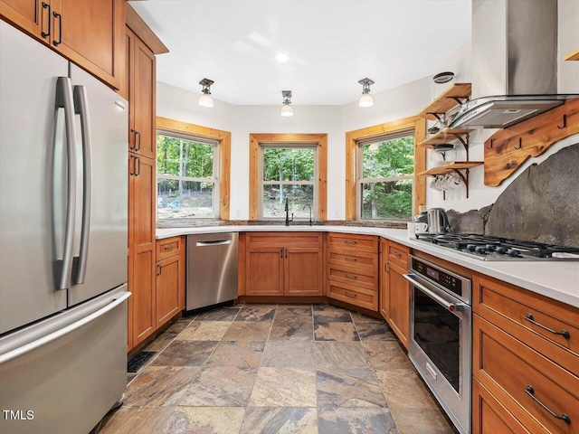 kitchen featuring wall chimney range hood, brown cabinets, appliances with stainless steel finishes, a sink, and stone finish floor
