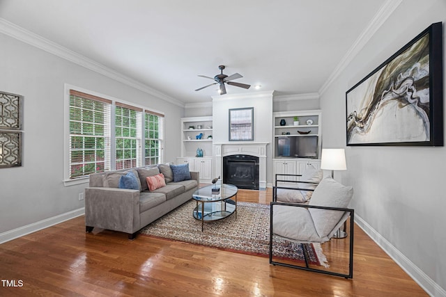 living area featuring a glass covered fireplace, a ceiling fan, baseboards, wood finished floors, and ornamental molding