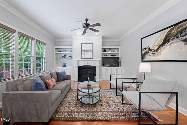 living room featuring a ceiling fan, crown molding, wood finished floors, baseboards, and a glass covered fireplace