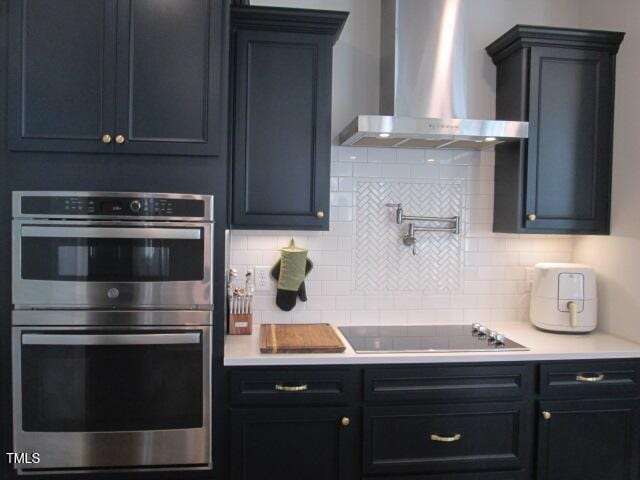 kitchen with decorative backsplash, double oven, black electric stovetop, wall chimney range hood, and light countertops