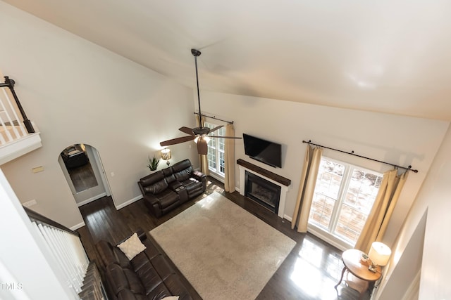 living area featuring lofted ceiling, a glass covered fireplace, ceiling fan, dark wood-style flooring, and baseboards