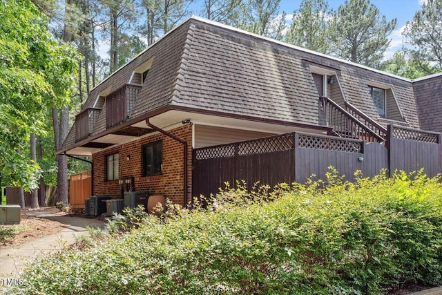 view of side of home featuring fence, roof with shingles, mansard roof, brick siding, and central AC unit
