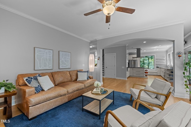 living area featuring a ceiling fan, crown molding, arched walkways, baseboards, and light wood finished floors