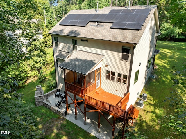 rear view of house featuring roof with shingles, a wooden deck, roof mounted solar panels, a lawn, and a patio
