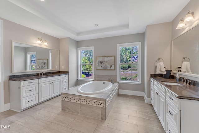 full bath featuring a tray ceiling, two vanities, a sink, and a bath