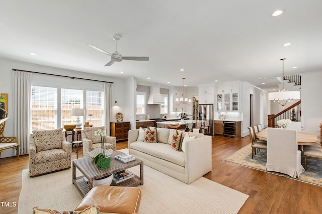 living area with light wood finished floors, ceiling fan with notable chandelier, stairs, recessed lighting, and wine cooler