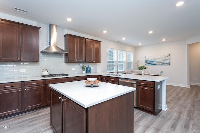 kitchen with a peninsula, wall chimney range hood, light wood-type flooring, a sink, and appliances with stainless steel finishes