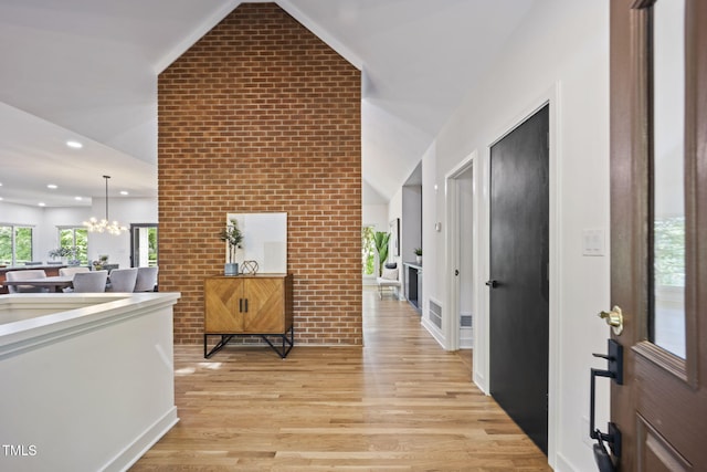 hallway with visible vents, an inviting chandelier, brick wall, light wood-style flooring, and vaulted ceiling