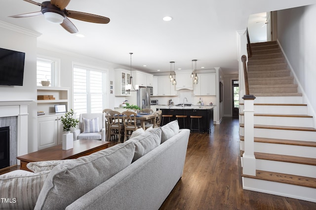 living area featuring a tiled fireplace, stairs, a ceiling fan, recessed lighting, and ornamental molding