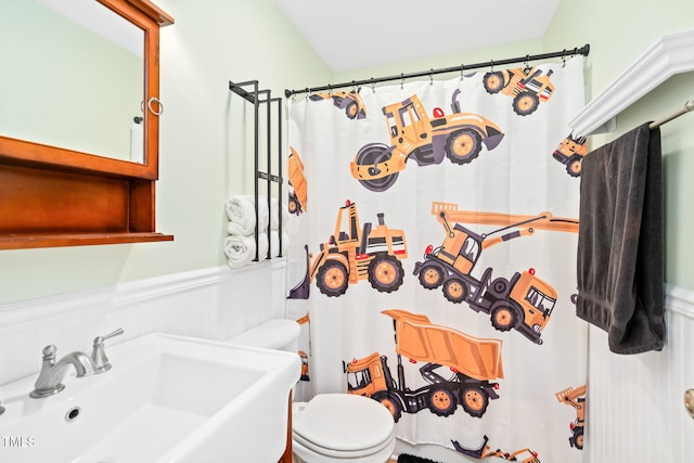 bathroom featuring wainscoting, a decorative wall, toilet, a shower with curtain, and a sink