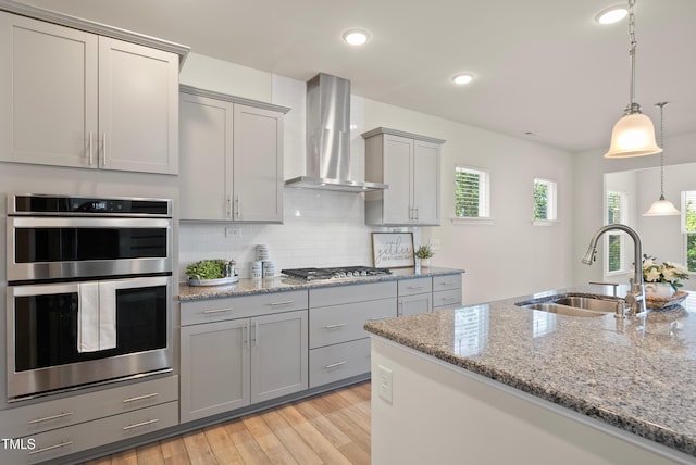 kitchen featuring wall chimney range hood, a sink, light wood-style flooring, backsplash, and gray cabinetry