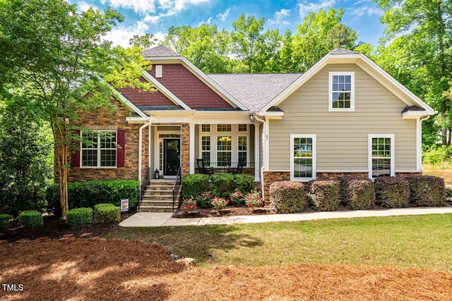 craftsman-style home with a front yard, roof with shingles, covered porch, and stone siding