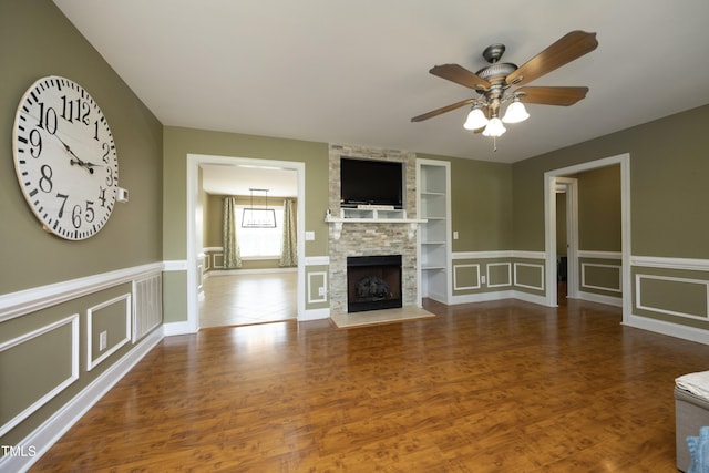 unfurnished living room with a decorative wall, wood finished floors, a stone fireplace, ceiling fan, and built in shelves