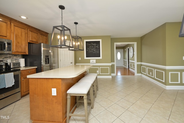 kitchen with a notable chandelier, appliances with stainless steel finishes, light countertops, brown cabinetry, and a kitchen bar