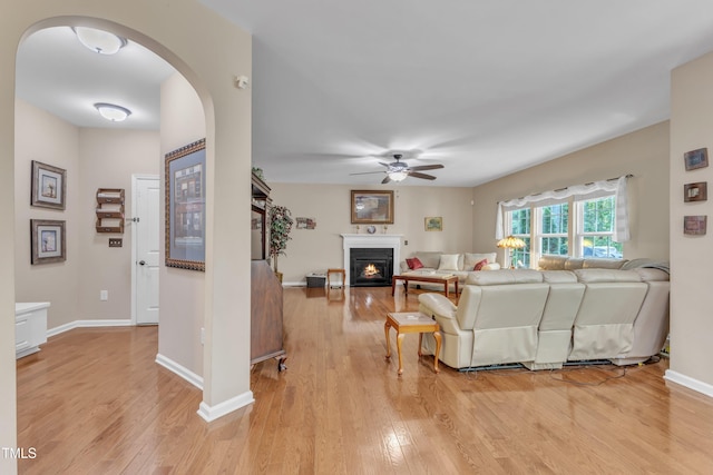 living room with arched walkways, a lit fireplace, a ceiling fan, and light wood finished floors