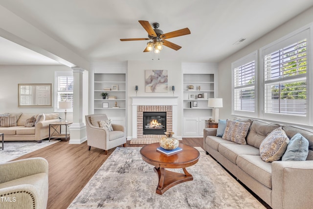 living area with decorative columns, a ceiling fan, a fireplace, built in shelves, and wood finished floors