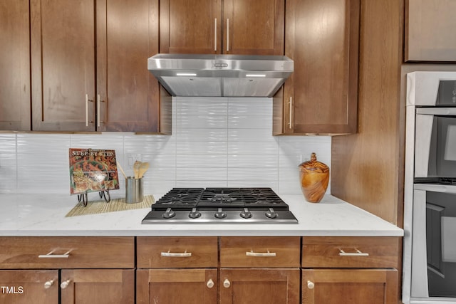 kitchen with brown cabinets, stainless steel gas cooktop, tasteful backsplash, under cabinet range hood, and multiple ovens