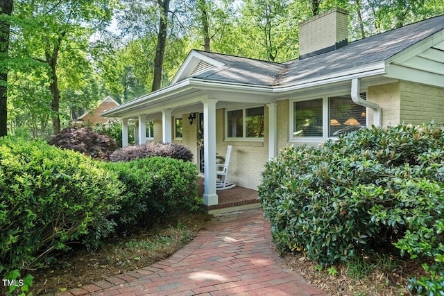 view of exterior entry with covered porch, brick siding, a chimney, and a shingled roof