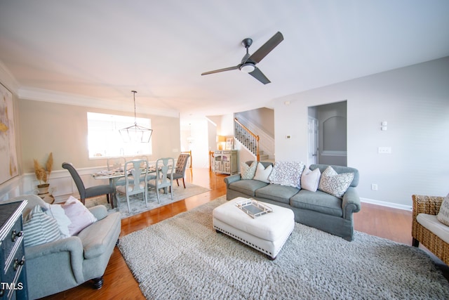 living room featuring ornamental molding, wood finished floors, baseboards, stairway, and ceiling fan with notable chandelier