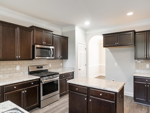 kitchen with light wood-style flooring, arched walkways, dark brown cabinetry, backsplash, and appliances with stainless steel finishes