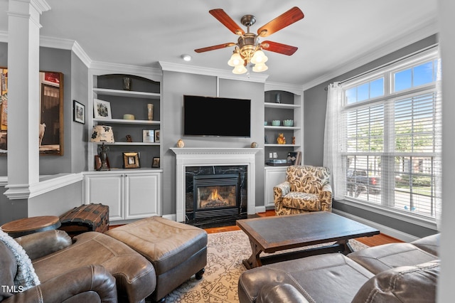 living area with crown molding, ceiling fan, ornate columns, built in shelves, and a premium fireplace