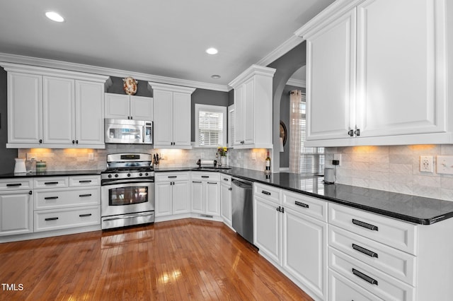kitchen featuring dark countertops, light wood-style floors, white cabinets, arched walkways, and appliances with stainless steel finishes